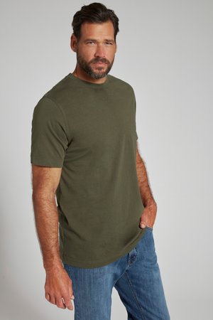 Mann mit Bart, der ein olivgrünes kurzärmeliges T-Shirt und blaue Jeans trägt, steht mit einer Hand in der Tasche vor einfarbigem Hintergrund.