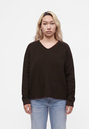 Jeune personne aux longs cheveux blonds portant un pull marron foncé et un jean bleu clair, debout devant un fond blanc uni.