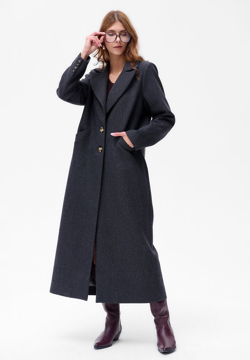 Lang, donkergrijze wolblendjas met een gelapte kraag, twee voorzakken en gouden knopen. Gepaard met bordeauxrode kniehoge laarzen.