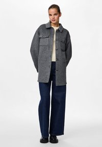 Γκρι πουκάμισο-τζάκετ από μαλλί με γιακά, δύο μπροστινές τσέπες και κουμπιά, φορώντας πάνω από μια κρεμ μπλούζα και μπλε παντελόνι με φαρδιές γραμμές.