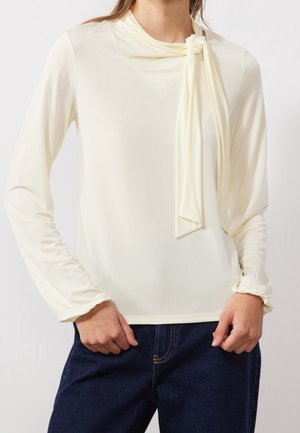 Vrouw in een crèmekleurige top met lange mouwen en een geknoopte sjaal detail op de schouder, in combinatie met donkerblauwe spijkerbroek.