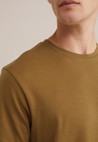 Alyvuogių žalia krūtinės apykaklės marškinėliai, pagaminti iš minkštos, lengvos medžiagos su trumpomis rankovėmis, pasižymintys tvarkingais siūlėmis ir klasikinio kirpimo.