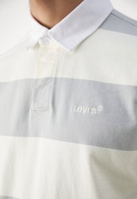 Personne portant un polo rugby Levi's rayé blanc et gris avec un col blanc, logo brodé sur la poitrine.