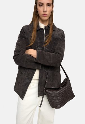 Młoda kobieta w ciemnej zamszowej kurtce, jasnym swetrze, białych spodniach, trzymająca tkany ciemny torebkę na ramię.