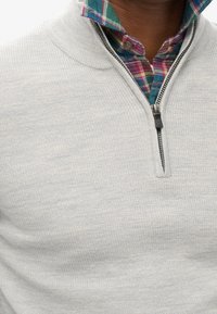 Pull gris à fermeture éclair avec un motif en maille texturée, porté par-dessus une chemise à carreaux bleue et verte avec des accents rouges et jaunes.