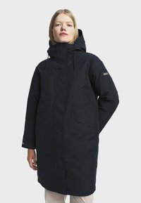 Femme portant un long manteau d'hiver noir avec capuche et poches avant, se tenant contre un fond clair.