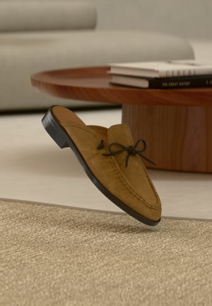 Mocassin à enfiler en daim marron avec nœud en cuir et semelle en caoutchouc noire, présentant des détails cousus et une texture douce.