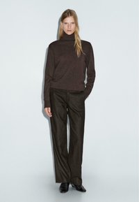 Pull en maille marron à col roulé associé à un pantalon large marron foncé et des chaussures noires à enfiler. Le modèle se tient devant un fond clair.