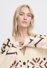 Cárdigan de punto crema con patrones geométricos en marrón, negro y rosa. Presenta un escote en V profundo y grandes botones marrones. Textura suave y ajuste relajado.