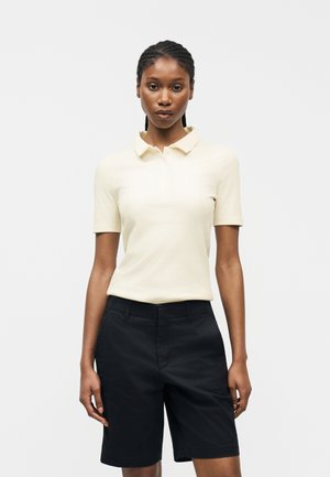 Vrouw met gevlochten haar, gekleed in een crème korte mouw polo en zwarte knielange shorts, staand tegen een effen witte achtergrond.