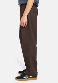 Brown, straight-leg pants with a smooth texture, a back pocket, and subtle stitching details, worn with black sneakers featuring a gum sole.