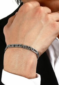 Silvermetallarmband med sammanlänkade geometriska länkar. Slät yta med polerad finish och subtil textur, visad på ett handled.