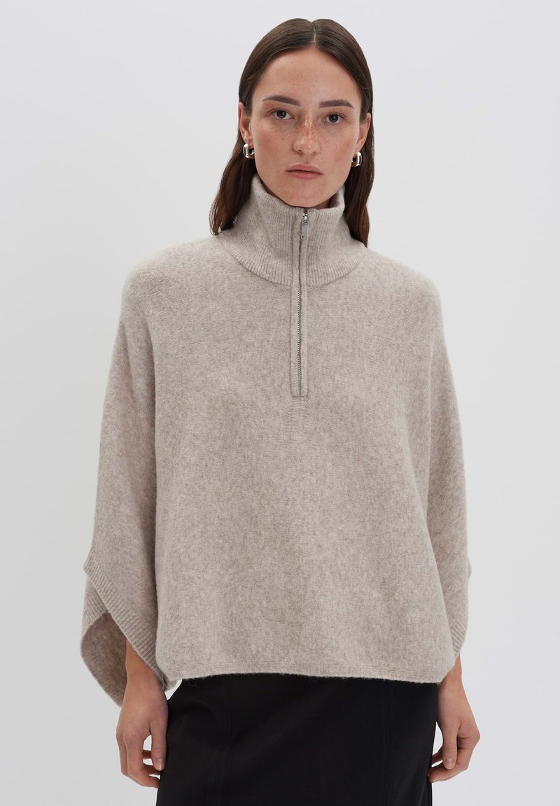 Woman with brown hair wears beige high-neck zippered poncho-style sweater and black skirt, standing against plain light background.