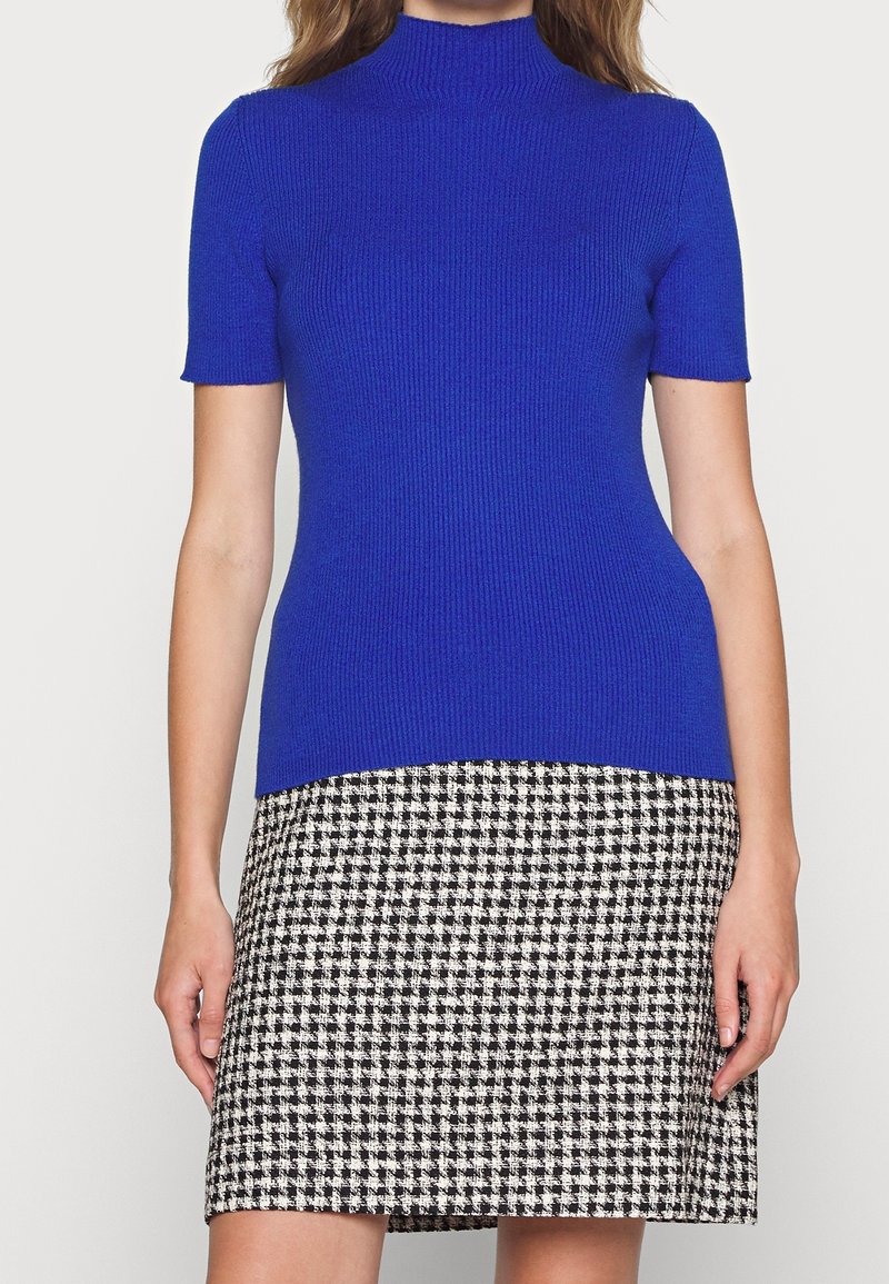 Femme portant un haut côtelé à manches courtes bleu vif associé à une jupe à motif pied-de-poule noir et blanc.