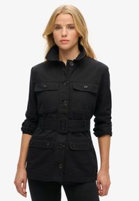 Chaqueta utilitaria negra hecha de tela de algodón. Presenta un cuello alto, cierre de botones, cuatro bolsillos frontales y una cintura con cinturón.