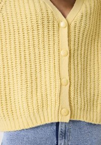 Cárdigan de punto amarillo con cuatro botones redondos, usado sobre unos jeans de denim azules con pequeños clavos plateados.