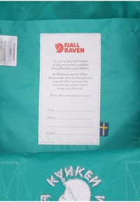 Mochila Kånken turquesa con un logo bordado en blanco, una etiqueta de tela que presenta las instrucciones de cuidado y una pequeña etiqueta de bandera sueca.