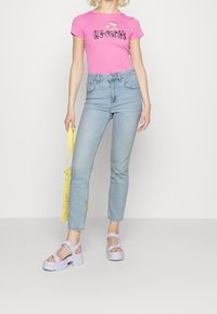Camiseta ajustada rosa con gráfico de "Wonder", jeans de tiro alto azul claro y sandalias gruesas lila; sosteniendo una bolsa de red amarilla.