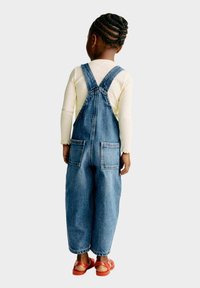 Denim tuinbroek met een losse pasvorm, voorzien van verstelbare bandjes, twee achterzakken en een vervaagde blauwe kleur. Gedragen over een licht, langemouwen shirt.