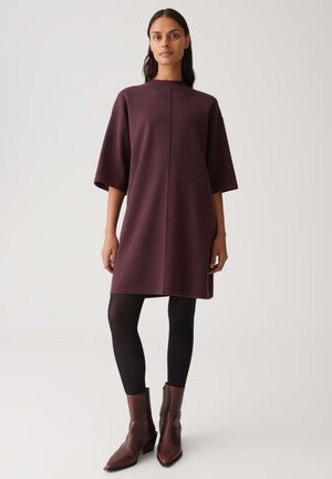 Eine Frau trägt ein lockeres, knielanges Kleid in Pflaumenfarbe mit Ellenbogen-langen Ärmeln, schwarze Strumpfhosen und braune Stiefeletten und steht vor einem schlichten Hintergrund.