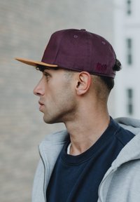 Cappello bordeaux con visiera marrone, con un logo a forma di conchiglia sul retro. Il portatore ha i capelli corti ed è vestito con una felpa grigia e una maglietta blu.