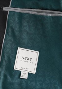 Copertura per abiti su misura verde con motivo a rombi, con etichetta bianca che dettaglia marca, vestibilità, taglia e paese di produzione.