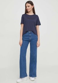 T-shirt in cotone blu navy con maniche corte e logo ricamato sul petto, abbinato a jeans in denim a vita alta e gamba larga e sneaker bianche.