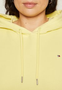 Zbliżenie na osobę noszącą jasnosłomkową bluzę z kapturem z sznurkami i małym prostokątnym logo na klatce piersiowej.