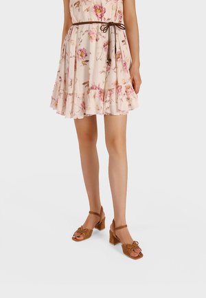 Donna che indossa un vestito corto rosa chiaro a fiori con una sottile cintura marrone e sandali marroni con tacco e cinturino alla caviglia, in piedi davanti a uno sfondo semplice.