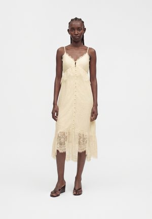 Femme avec des tresses portant une robe midi en dentelle crème sans manches avec fermeture boutonnée sur le devant et ourlet en dentelle transparente, debout sur fond blanc.