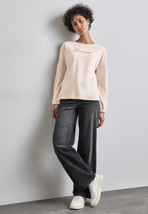 Hellbeige Sweatshirt mit umgeschlagenen Ärmeln und besticktem Text. Kombiniert mit dunkelgrauen weit geschnittenen Jeans und weißen Sneakers.