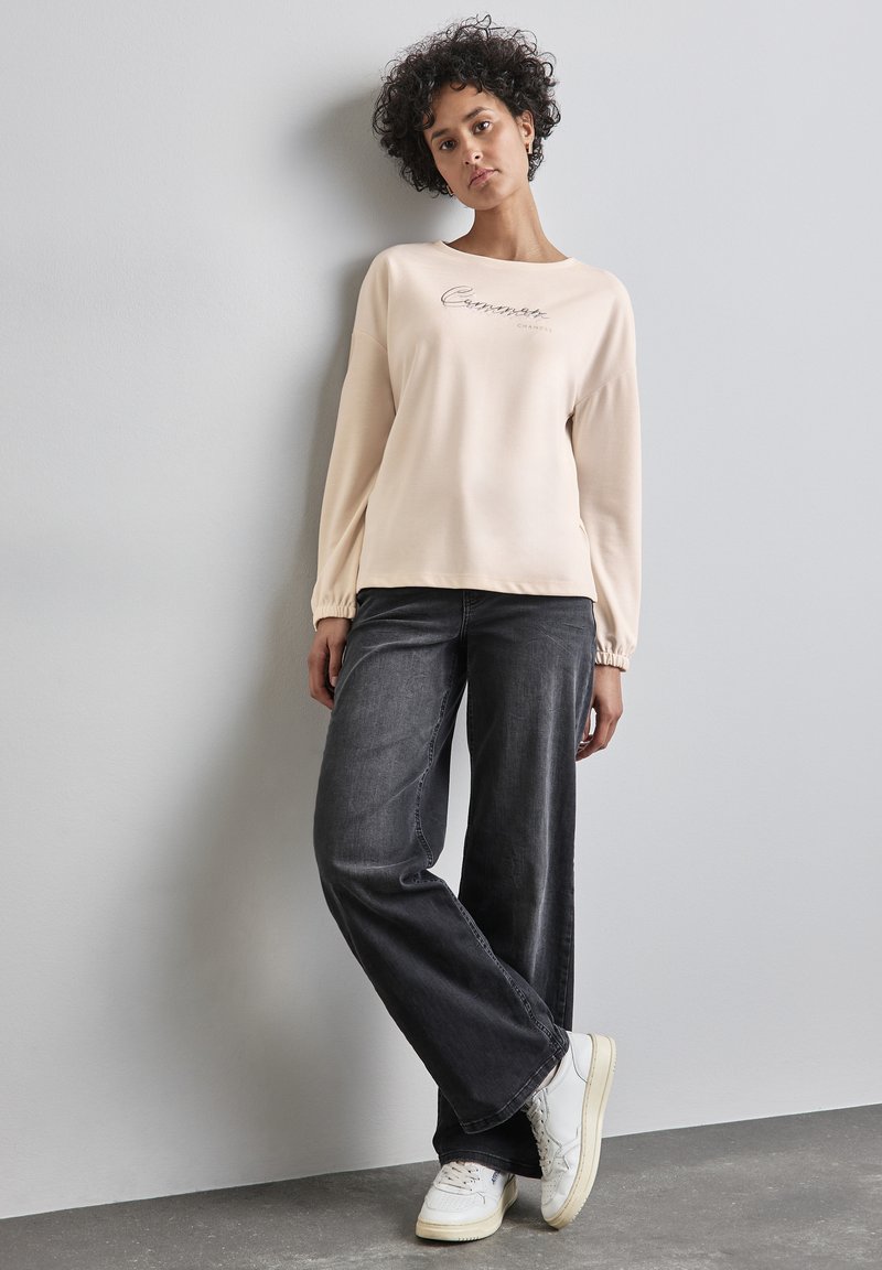 Hellbeige Sweatshirt mit umgeschlagenen Ärmeln und besticktem Text. Kombiniert mit dunkelgrauen weit geschnittenen Jeans und weißen Sneakers.