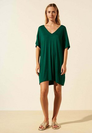 Robe verte en V faite d'un tissu doux, avec des manches larges, une coupe ample et un ourlet qui tombe au-dessus du genou. Portée avec des sandales dorées.