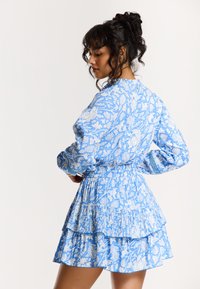 Robe florale bleue avec des manches longues, un ourlet froncé à volants et une taille ajustée. Le tissu a une texture lisse avec des motifs blancs intricats.