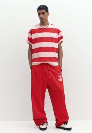 Giovane uomo con pantaloni rossi larghi e una maglietta a righe rosse e rosa, in piedi con le mani in tasca su sfondo bianco.