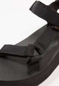 Sandale noire avec semelle en caoutchouc texturée, dotée de brides en nylon ajustables. Détail de nœud mis en valeur à l'intersection des brides.