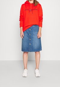 Felpa rossa con cappuccio e lacci, abbinata a una gonna di jeans blu con chiusura a bottoni frontale e sneakers bianche.