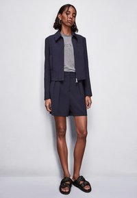 Giacca cropped blu navy con colletto, abbinata a pantaloni shorts plissettati dello stesso colore. T-shirt grigia sotto e sandali neri annodati. Tessuto dalla texture liscia.
