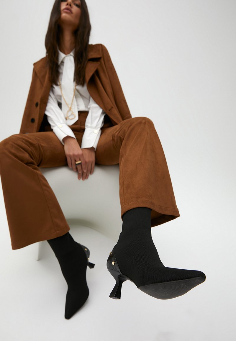 Černá ponožková bota s špičatou špičkou a tenkým podpatkem. Obsahuje zlaté akcenty, umístěné na minimalistickém pozadí.