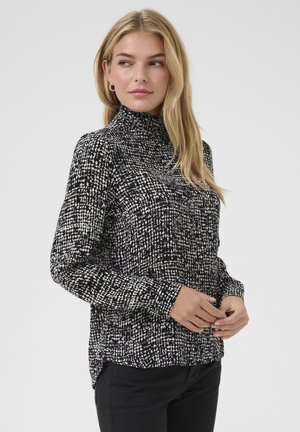 Haut à col roulé présentant un motif tacheté noir et blanc, des manches longues, et une coupe décontractée avec un ourlet arrière légèrement plus long.