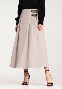 Beżowa plisowana spódnica z wysokim paskiem, wyposażona w dwa brązowe skórzane paski z srebrnymi klamrami, wykonana z gładkiej tkaniny, sięgająca do podłogi.