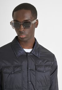 Giacca imbottita blu navy con un ampio colletto, doppie tasche sul petto e chiusure a bottoni neri; abbinata a occhiali da sole geometrici con montatura trasparente.
