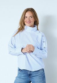 Randig blåvit topp med hög krage, långa ärmar och knappdetaljer på ärmarna, bärs över ljusblå jeans.