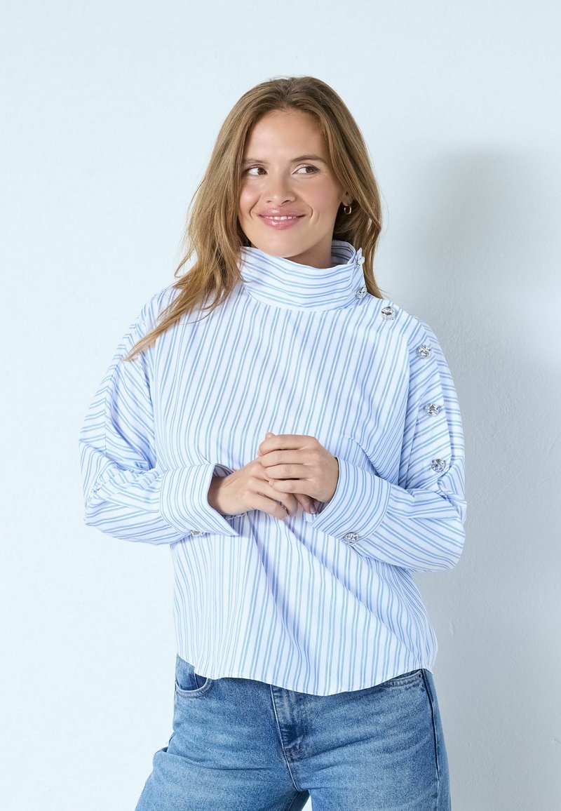 Randig blåvit topp med hög krage, långa ärmar och knappdetaljer på ärmarna, bärs över ljusblå jeans.