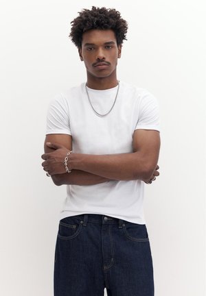 Junger Mann mit lockigem Haar, der ein weißes T-Shirt, eine silberne Halskette, ein Armband und dunkelblaue Jeans trägt, steht mit verschränkten Armen.