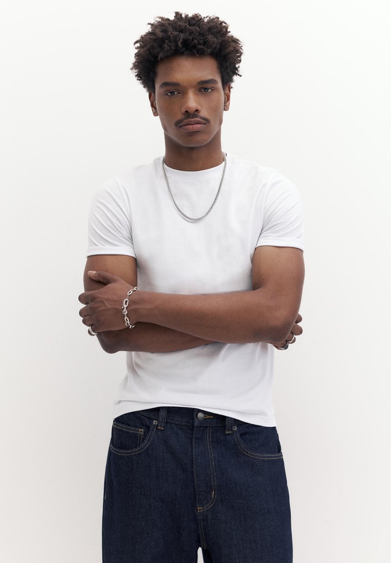 Ung mand med krøllet hår iført hvid t-shirt, sølvkæde, armbånd og mørkeblå jeans, stående med armene over kors.