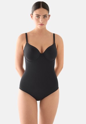 Mujer joven con cabello oscuro recogido, vestida con un body negro ajustado de tirantes finos, de pie contra un fondo claro y liso.