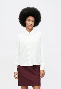 Woman with large afro hairstyle wearing a white blouse with pearl-trimmed collar and a burgundy skirt against a plain background.
