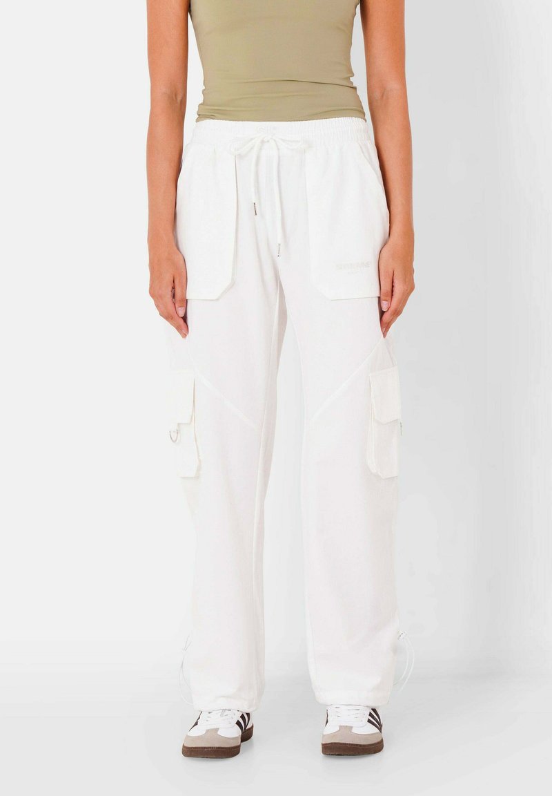 Persoon in witte cargobroek met voor- en zijzakken, beige tanktop en witte sneakers met bruine zolen.