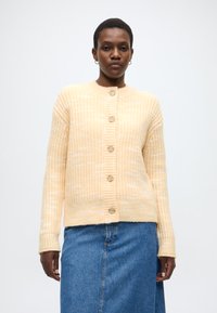 Καρντιγκάν με κουμπιά σε απαλό κίτρινο πλεκτό, με ριμπ ζώνη και μανσέτες, με πέντε ξύλινα κουμπιά στο μπροστινό μέρος, φορεμένο πάνω από φούστα από τζιν.